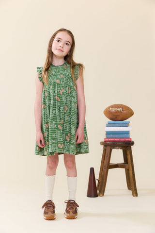 Pink Chicken Girls Stevie Dress - Football Field
A young girl stands in a studio wearing a green sleeveless dress with a brown and white abstract pattern. She is styled with white socks and brown sneakers, standing next to a small wooden stool holding a stack of books and a football on a beige background.
