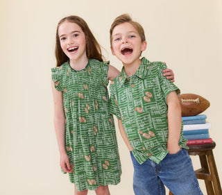 Pink Chicken Girls Stevie Dress - Football Field
A girl and a boy wearing matching green football-field print outfits, standing together against a plain beige background. The girl wears a short-sleeve ruffle-shoulder dress, and the boy wears a short-sleeve collared shirt with jeans.
