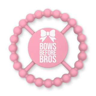 Bella Tunno Happy Teether - Bows Before Bros
A pink silicone baby teether with a circular beaded outer ring and a central round pad that reads 'BOWS BEFORE BROS' in white text with a bow icon above. The brand name 'Della Ferraro' is subtly printed along the bottom edge of the center pad.