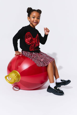 Lola and The Boys Big Bow Sweater
Child sitting on a large red ball with a pacifier attached, wearing a black sweater with a red sequin bow and a patterned skirt.