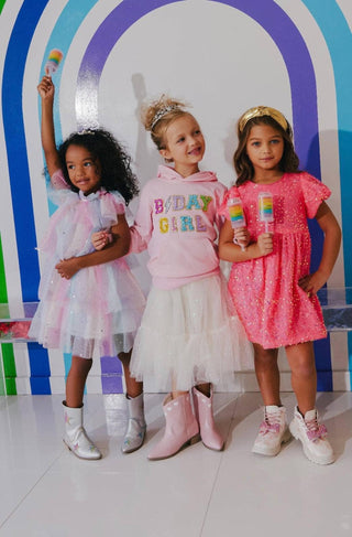 Lola and The Boys Birthday Girl Hoodie
Three children in colorful outfits standing in front of a rainbow arch.