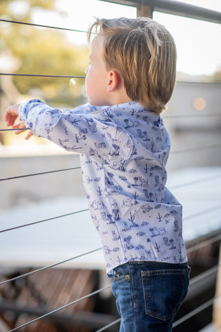 BlueQuail Clothing Co Long Sleeve Shirt - Terlingua Ranch
A young boy with blonde hair stands by a modern cable railing, wearing a light blue long-sleeve button-up shirt with a dark blue nautical print and dark blue jeans.