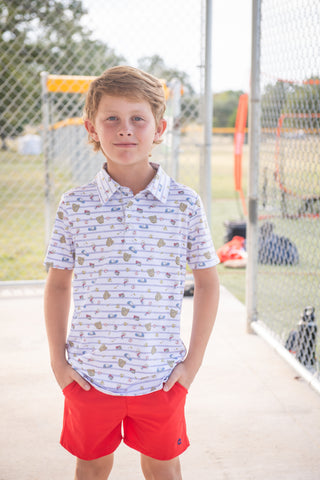 BlueQuail Clothing Co Polo Short Sleeve Shirt - 7th Inning Stretch
Young boy wearing a patterned shirt and red shorts standing in front of a chain-link fence.