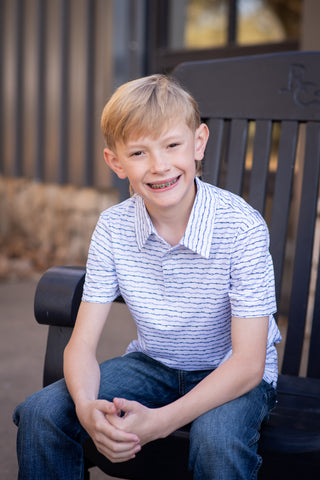 BlueQuail Clothing Co Polo Short Sleeve Shirt - Barbwire
A young blond boy with braces sits smiling on a black outdoor bench, wearing a white short-sleeve polo shirt with thin blue horizontal stripes and blue jeans.