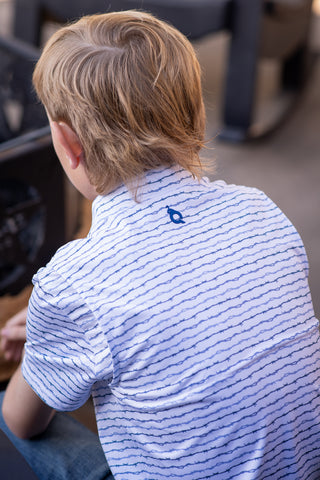 BlueQuail Clothing Co Polo Short Sleeve Shirt - Barbwire
A child viewed from behind wearing a white short-sleeve polo shirt with thin dark horizontal stripes and a small blue logo on the upper back, seated in a real environment with a blurred background.