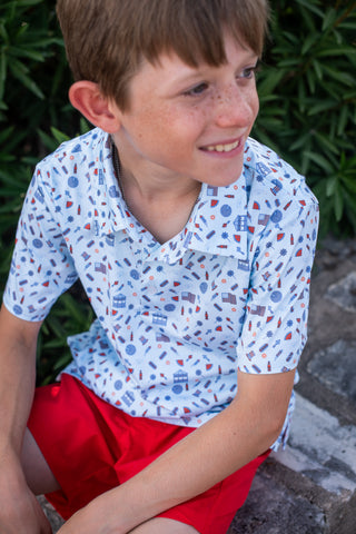 BlueQuail Clothing Co Polo Short Sleeve Shirt - Stars & Stripes
Young boy wearing a patterned shirt and red shorts sitting outdoors.