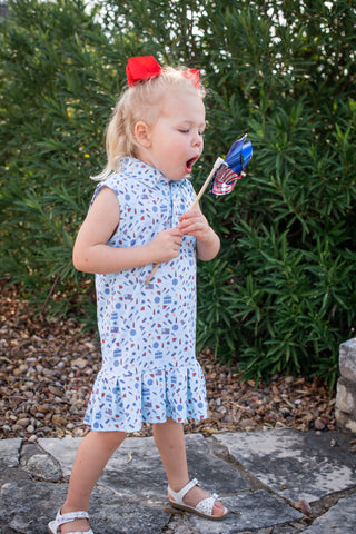 BlueQuail Clothing Co Polo Sleeveless Dress - Stars & Stripes
Child in a floral dress holding a colorful toy outdoors with greenery in the background
