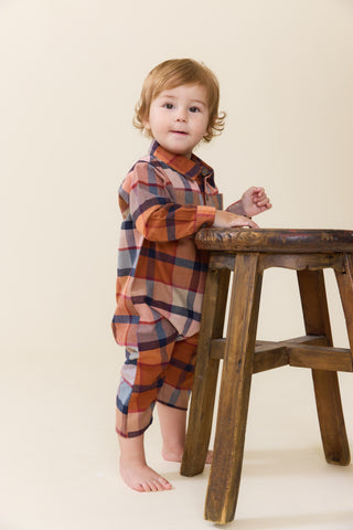 Blue Rooster Baby Boys Easton Jumper - Hayride Plaid
A toddler standing barefoot next to a rustic wooden stool, wearing a long-sleeve plaid shirt and matching pants in rust orange, navy blue, and white.