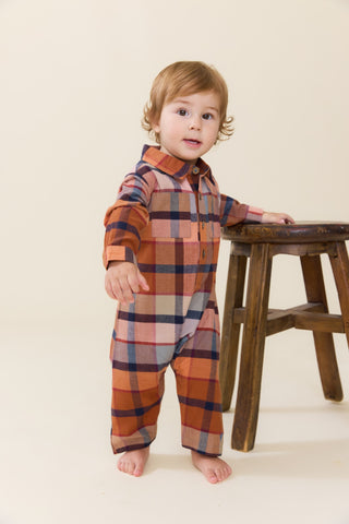 Blue Rooster Baby Boys Easton Jumper - Hayride Plaid
A toddler standing barefoot next to a wooden stool, wearing a long-sleeve plaid jumpsuit in rust orange, navy blue, and beige tones against a warm cream background.