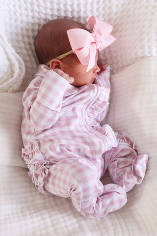 Caden Lane Peter Pan Collar Ruffle Zipper Footie - Rosy Gingham
Newborn baby in a pink checkered outfit with a large bow on a white blanket
