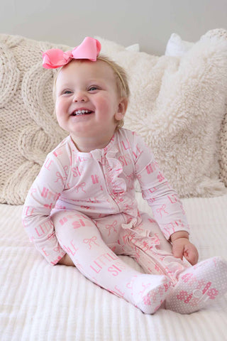 Caden Lane Ruffle Zipper Footie - Little Sis Bows
Baby in pink pajamas with a bow sitting on a white couch.