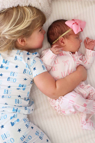 Caden Lane Two Piece Pajama Set - Big Bro Stars
Child wearing 'Big Bro' pajamas holding a baby with a pink bow, both lying on a soft surface.