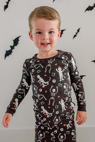 Caden Lane Two Piece Pajama Set - Glow in the Dark Eye Want Candy A young boy wearing a long-sleeve dark brown pajama shirt with a white skeleton and Halloween-themed print, standing against a white wall decorated with black bat cutouts.
