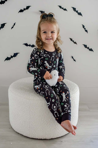 Caden Lane Two Piece Pajama Set - Glow in the Dark Eye Want Candy
A young girl with blonde hair in a high ponytail sits on a round white pouf, wearing a black long-sleeve pajama set with a white skeleton and ghost pattern. She is holding a white plush ghost toy against a white wall decorated with black bat cutouts.