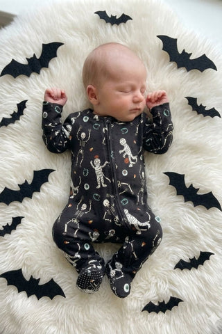 Caden Lane Zipper Footie - Glow in the Dark Eye Want Candy
A sleeping newborn baby lies on a white faux-fur blanket surrounded by black bat cutouts, wearing a black footed one-piece outfit with a white skeleton and ghost print.