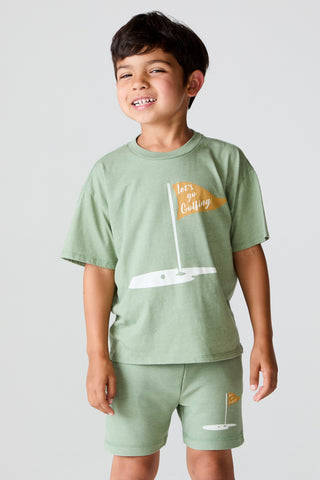 Chaser On The Green Tee
Child wearing a green t-shirt and shorts set with a sailboat graphic on a light gray background