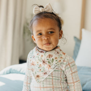 Copper Pearl Natalie Bandana Bib Set Young child wearing a floral and plaid outfit with a bow in their hair, sitting on a light blue cushion.
