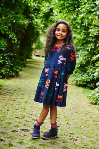 Pink Chicken Girls Brooke Dress - Multi Bow Applique
Young girl in a colorful dress standing on a brick path with greenery around