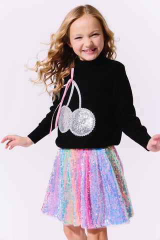 Lola and The Boys Crystal Cherry Sweater
Young girl wearing a black sweater with cherry design and a colorful sequin skirt on a white background