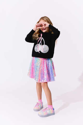 Lola and The Boys Crystal Cherry Sweater
Young girl wearing a black sweater with a graphic design and a colorful skirt on a white background