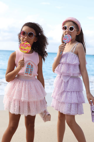 Lola and The Boys Crystal Chill Out Tank Top
Two young girls in pink dresses holding lollipop toys on a beach.