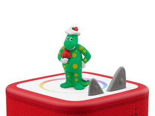 Tonies Character - The Wiggles: Dorothy The Dinosaur
Toy figure of a green dinosaur-like character with a red hat and flower, standing on a red toy box with shark design.