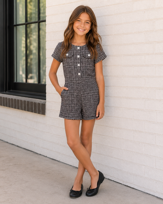 FBZ Black & Pink Tweed Romper
A young girl stands against a white brick wall wearing a short-sleeve tweed romper in a black and white micro-check pattern with white decorative buttons down the front. She is wearing black flats.