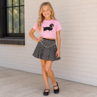 FBZ Dog on Pink Jersey Boxy Tee
A young girl stands against a white brick wall wearing a pink short-sleeve crop top with a black dachshund graphic, a black and white tweed mini skirt with a silver chain detail, and black Mary Jane shoes.