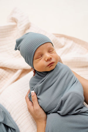Lou Lou and Company Ribbed Swaddle Blanket - Ford
Newborn baby wrapped in a blue blanket and wearing a matching blue hat on a soft pink blanket.