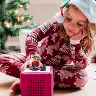 Tonies Character: Favorite Children’s Songs: Holiday Songs 2
Child in festive pajamas playing with a pink toy near a Christmas tree.