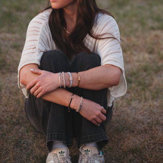 K'lani "Flirt" Polka Dot in Cream and Black Hair Tie Bracelets
A person sitting outdoors on dry grass wearing multiple delicate bracelets on both wrists, including silver chain bracelets, a beaded bracelet, and a gold-tone bracelet. They are also wearing a cream ribbed knit top, dark jeans, and white sneakers.