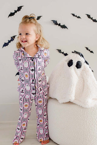 Caden Lane Girl's Two Piece Flare Lounge Set - Glow in the Dark Galloween
A young girl stands barefoot next to a large white plush ghost, wearing a pink and white Halloween-themed pajama set with bat and pumpkin prints. The background is a light wall decorated with black bat cutouts.