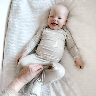 Gunamuna Convertible Romper Footie - Goose
Baby lying on a white blanket wearing a beige outfit with small patterns.