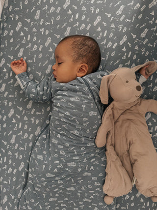 Gunamuna Pup Lovey - Mutts
A sleeping baby wearing a gray long-sleeve swaddle blanket with a white pattern, lying next to a beige plush toy on a matching patterned blanket.