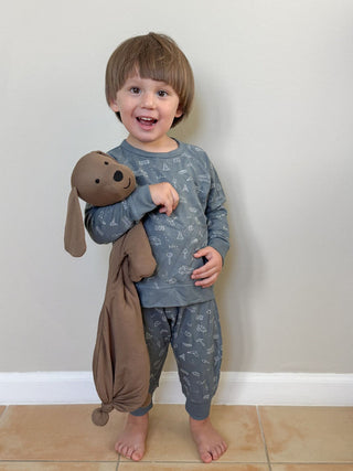 Gunamuna Pup Lovey - Peanut
Child wearing a gray pajama set with a brown dog-shaped blanket against a plain wall.