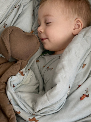 Gunamuna Pup Lovey - Peanut
Baby wrapped in a gray blanket with animal patterns, lying next to a brown plush toy.