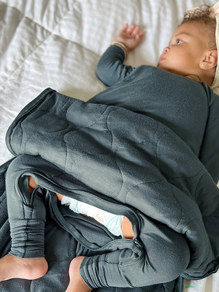 Gunamuna Sleep Sack 1.0 TOG - Hush
A baby wearing a dark slate blue long-sleeve outfit and diaper cover, lying on a white quilted bed and wrapped in a matching dark slate quilted blanket.