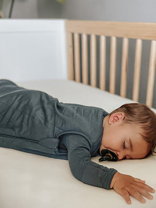 Gunamuna Sleep Sack 1.0 TOG - Hush
A baby sleeping soundly in a dark slate blue long-sleeve swaddle or sleep sack, lying on their side on a white mattress next to a wooden crib.