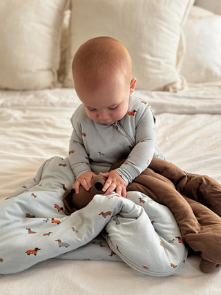 Gunamuna Sleep Sack 1.0 TOG - Woof
Baby sitting on a bed with a gray blanket and brown pants