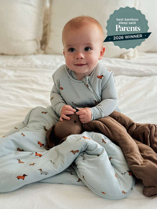 Gunamuna Sleep Sack 1.0 TOG - Woof
A baby sitting on a white bed wearing a light blue long-sleeve sleep sack with a brown dachshund print, wrapped in a brown plush blanket. A teal badge in the top right reads 'best bamboo sleep sack Parents 2026 WINNER'.