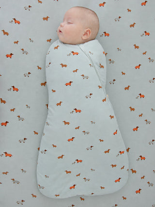 Gunamuna Swaddle Sleep Sack 1.0 TOG - Woof
A sleeping newborn baby swaddled in a pale blue swaddle blanket with a repeating pattern of small orange and gray dachshunds, lying on a matching patterned sheet.