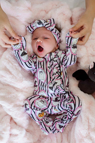 Caden Lane Ruffle Zipper Footie - Glow in the Dark Galloween
A baby wearing a black and white zebra-print footed sleeper with pink heart outlines and cartoon cat faces, along with a matching headband, lying on a pink plush blanket with hands holding the baby's hands.