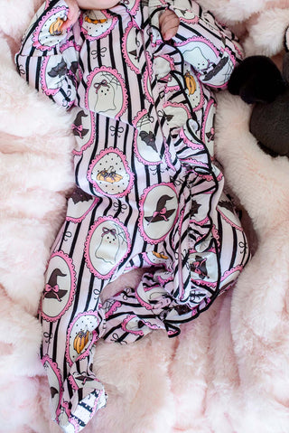 Caden Lane Ruffle Zipper Footie - Glow in the Dark Galloween
A baby's footed sleep suit with a Halloween print, featuring black and white stripes, pink-trimmed ghost frames, and witch hats, laid out on a soft pink faux fur blanket.