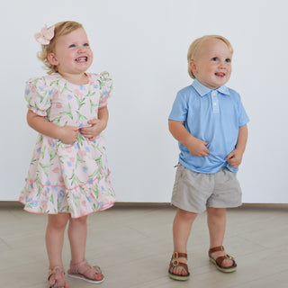 Hollis Baker Everly Dress - Tulips
Two children, a girl in a floral dress and a boy in a blue shirt and shorts, standing on a light-colored floor.