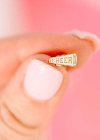 Pierced Co. Cheer Megaphone Earrings
Gold 'CHEER' pin on a hand with a pink background