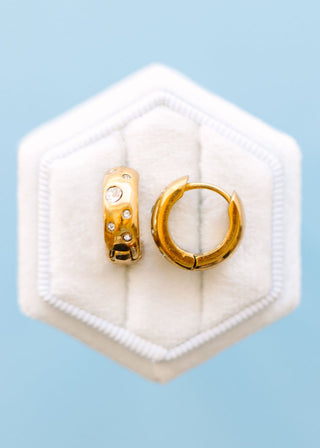 Pierced Co. Huggies - Diamond Dot
A pair of gold hoop earrings, one with small clear stones, displayed on a white hexagonal velvet jewelry stand against a light blue background.