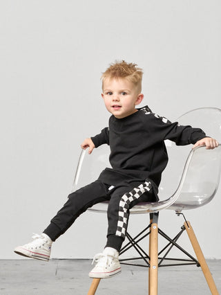 Little Bipsy Stripe Checkered Jogger - Black
Child wearing a black outfit with white text and checkered details, sitting on a clear chair against a gray background.