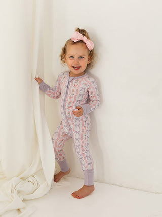 In My Jammers Zipper Romper - Purple Amelia Bow
Child wearing a patterned onesie standing against a white curtain.