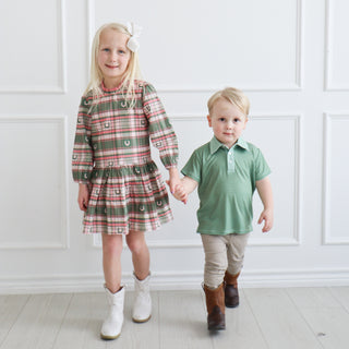 Hollis Baker Trey Polo - Green Stripe
A young girl and a young boy walking hand in hand in a bright room with white paneled walls and light wood floors. The girl wears a long-sleeve plaid dress in red, green, and beige with white boots, and the boy wears a green polo shirt, beige pants, and brown boots.