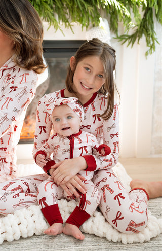 In My Jammers Merry Little Bows Zipper Romper
Two children in matching pajamas sitting on a rug with a fireplace and Christmas tree in the background.